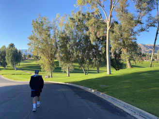 A sunlit outdoor scene with a person wearing a red capella cap, smiling as they walk through a park.