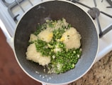 In a dark, non-stick frying pan on a stovetop, there is a mixture of uncooked white rice, green peas, chopped spring onions, and minced garlic. The pan is placed on a granite countertop beside a stove with visible burners.