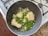 In a dark, non-stick frying pan on a stovetop, there is a mixture of uncooked white rice, green peas, chopped spring onions, and minced garlic. The pan is placed on a granite countertop beside a stove with visible burners.
