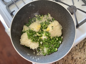 In a dark, non-stick frying pan on a stovetop, there is a mixture of uncooked white rice, green peas, chopped spring onions, and minced garlic. The pan is placed on a granite countertop beside a stove with visible burners.