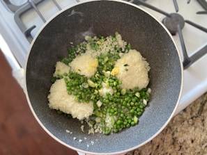In a dark, non-stick frying pan on a stovetop, there is a mixture of uncooked white rice, green peas, chopped spring onions, and minced garlic. The pan is placed on a granite countertop beside a stove with visible burners.