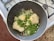 In a dark, non-stick frying pan on a stovetop, there is a mixture of uncooked white rice, green peas, chopped spring onions, and minced garlic. The pan is placed on a granite countertop beside a stove with visible burners.