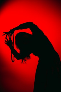 A silhouette of a person holding a string of beads against an intense red background. The figure appears to be in motion, with arms bent and fingers splayed, creating a dynamic and artistic pose.