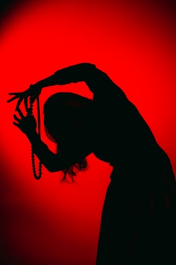 A silhouette of a person holding a string of beads against an intense red background. The figure appears to be in motion, with arms bent and fingers splayed, creating a dynamic and artistic pose.