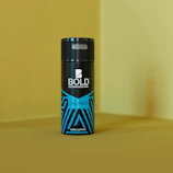 A can of deodorant, labeled BOLD, stands upright against a yellow background. The bottle features a sleek black design with blue geometric patterns and the word ALPHA written on it.