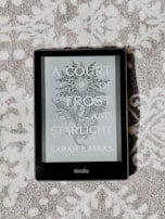 A Kindle device displaying the book cover of 'A Court of Frost and Starlight' by Sarah J. Maas is lying on a textured, patterned surface. The cover features a decorative floral illustration in grayscale.