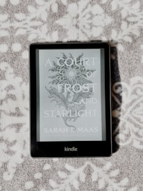 A Kindle device displaying the book cover of 'A Court of Frost and Starlight' by Sarah J. Maas is lying on a textured, patterned surface. The cover features a decorative floral illustration in grayscale.