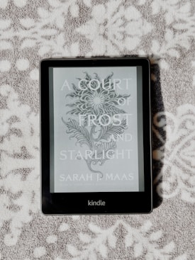 A Kindle device displaying the book cover of 'A Court of Frost and Starlight' by Sarah J. Maas is lying on a textured, patterned surface. The cover features a decorative floral illustration in grayscale.