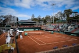 A tennis court with clay surface surrounded by stands, partially filled with spectators. Several chairs are positioned courtside, with some staff present. Banners and advertisements are visible around the court area. The sky above is partly cloudy with patches of blue.