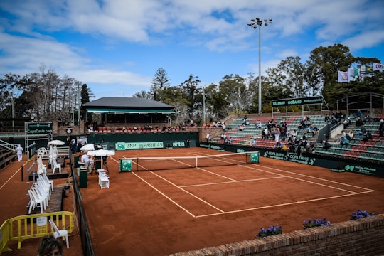 A tennis court with clay surface surrounded by stands, partially filled with spectators. Several chairs are positioned courtside, with some staff present. Banners and advertisements are visible around the court area. The sky above is partly cloudy with patches of blue.