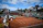 A tennis court with clay surface surrounded by stands, partially filled with spectators. Several chairs are positioned courtside, with some staff present. Banners and advertisements are visible around the court area. The sky above is partly cloudy with patches of blue.