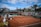 A tennis court with clay surface surrounded by stands, partially filled with spectators. Several chairs are positioned courtside, with some staff present. Banners and advertisements are visible around the court area. The sky above is partly cloudy with patches of blue.