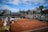 A tennis court with clay surface surrounded by stands, partially filled with spectators. Several chairs are positioned courtside, with some staff present. Banners and advertisements are visible around the court area. The sky above is partly cloudy with patches of blue.