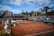 A tennis court with clay surface surrounded by stands, partially filled with spectators. Several chairs are positioned courtside, with some staff present. Banners and advertisements are visible around the court area. The sky above is partly cloudy with patches of blue.