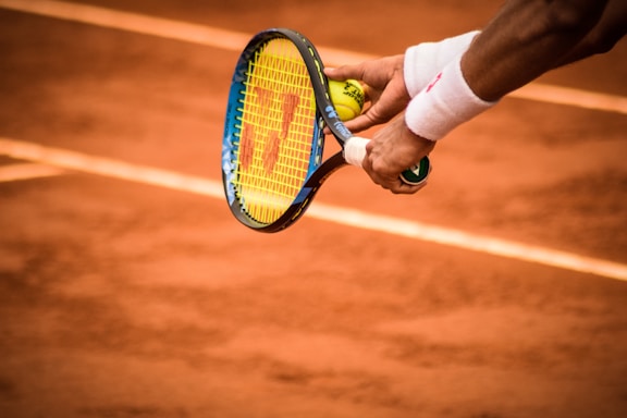 A close-up of a tennis player preparing to serve, holding a racket and a tennis ball. The court is clay, and the player's wristbands and racquet details are visible.