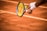 A close-up of a tennis player preparing to serve, holding a racket and a tennis ball. The court is clay, and the player's wristbands and racquet details are visible.