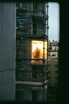 A construction site with partially completed scaffolding surrounds a building. A brightly lit room reveals a person standing inside, creating a stark contrast with the dark exterior. The lower-light setting adds to the mood, capturing a moment during either dusk or dawn. Adjacent buildings are visible in the background, indicating an urban environment.