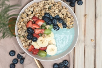 A healthy breakfast bowl filled with granola, a mix of fresh fruits including bananas, strawberries, blueberries, and green grapes, topped on a base of creamy yogurt with a hint of green color. The bowl is placed on a soft beige cloth beside scattered blueberries and a wooden surface.
