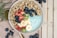 A healthy breakfast bowl filled with granola, a mix of fresh fruits including bananas, strawberries, blueberries, and green grapes, topped on a base of creamy yogurt with a hint of green color. The bowl is placed on a soft beige cloth beside scattered blueberries and a wooden surface.