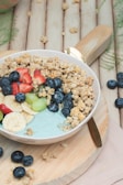 A bowl filled with a mixture of granola, fresh fruits including strawberries, bananas, blueberries, and grapes, set on a wooden board. The background features a rustic, wooden surface with some scattered granola and blueberries.