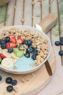 A bowl filled with a mixture of granola, fresh fruits including strawberries, bananas, blueberries, and grapes, set on a wooden board. The background features a rustic, wooden surface with some scattered granola and blueberries.