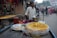 A street vendor stands behind a wooden cart filled with a large tray of golden fried snacks, likely pakoras, topped with green herbs. He is positioned in a bustling market street, surrounded by various shops and stands displaying different goods. People are walking and cycling in the background, hinting at a vibrant and busy atmosphere.