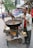 A street vendor is cooking snacks in a large wok over an open flame on a makeshift cart. He is stirring the food with a spatula. There are various items on the cart, such as a block of raw material and some containers. In the background, there are signs in a local language and a few people walking by. Trees and buildings are visible, suggesting a busy street environment.