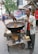 A street vendor is cooking snacks in a large wok over an open flame on a makeshift cart. He is stirring the food with a spatula. There are various items on the cart, such as a block of raw material and some containers. In the background, there are signs in a local language and a few people walking by. Trees and buildings are visible, suggesting a busy street environment.