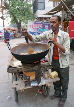 A street vendor is cooking snacks in a large wok over an open flame on a makeshift cart. He is stirring the food with a spatula. There are various items on the cart, such as a block of raw material and some containers. In the background, there are signs in a local language and a few people walking by. Trees and buildings are visible, suggesting a busy street environment.