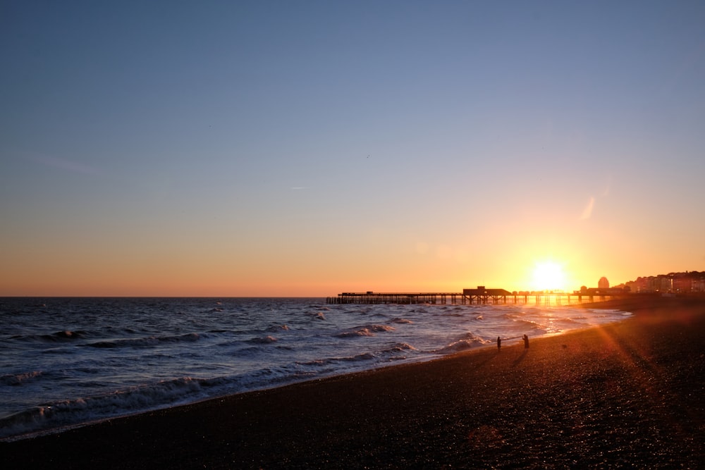 the sun is setting over the ocean with a pier in the distance