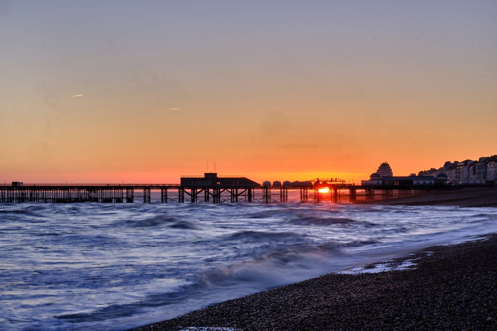 the sun is setting over the ocean with a pier in the background