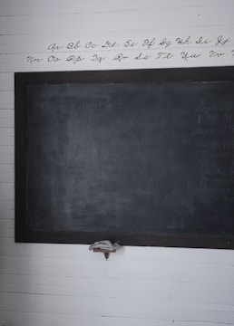 A wooden framed blackboard is mounted on a white, paneled wall. Above the blackboard, the alphabet is neatly written in cursive script. The blackboard appears slightly worn, with faint writing visible. A small shelf below holds an eraser.