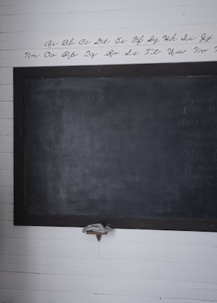A wooden framed blackboard is mounted on a white, paneled wall. Above the blackboard, the alphabet is neatly written in cursive script. The blackboard appears slightly worn, with faint writing visible. A small shelf below holds an eraser.