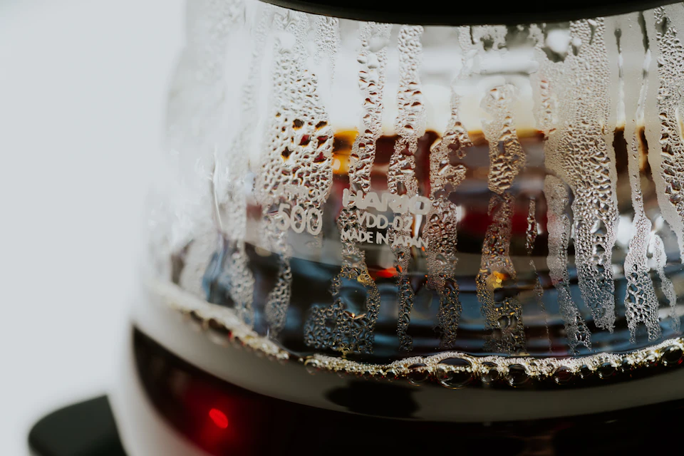 The image shows a close-up view of a coffee maker, specifically focusing on its glass carafe and water reservoir. The carafe is filled with dark brown liquid, possibly coffee or tea, which has formed droplets due to condensation from the hot beverage. The carafe features a label that reads "Dodmekar" in white text against a black background.