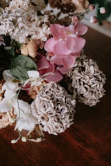 A close-up image of a bouquet consisting of various flowers. The flowers include pink orchids, withered hydrangeas, and some white blossoms. The petals are textured and dried, indicating the passage of time. The background is a dark wooden surface that emphasizes the flowers' details.