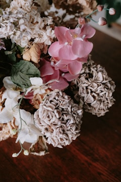 A close-up image of a bouquet consisting of various flowers. The flowers include pink orchids, withered hydrangeas, and some white blossoms. The petals are textured and dried, indicating the passage of time. The background is a dark wooden surface that emphasizes the flowers' details.