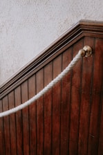 A close-up view of a dark wooden staircase with vertical paneling. There is a thick white rope handrail attached to a brass fitting on the wood, giving a classic or vintage look. The texture of the wall above the staircase is rough and light-colored, contrasting with the dark wood.