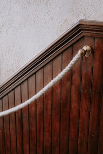 A close-up view of a dark wooden staircase with vertical paneling. There is a thick white rope handrail attached to a brass fitting on the wood, giving a classic or vintage look. The texture of the wall above the staircase is rough and light-colored, contrasting with the dark wood.