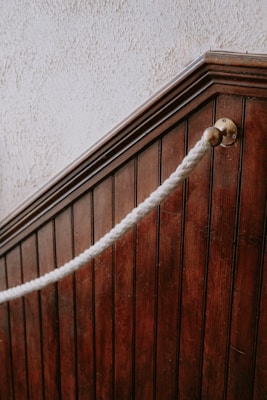 A close-up view of a dark wooden staircase with vertical paneling. There is a thick white rope handrail attached to a brass fitting on the wood, giving a classic or vintage look. The texture of the wall above the staircase is rough and light-colored, contrasting with the dark wood.