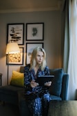 A person wearing dark blue pajamas with white star patterns is sitting on a comfortable sofa, holding a tablet. The room is softly lit by a nearby floor lamp, with framed artwork adorning the wall behind them. A green cushion adds a touch of color to the sofa.