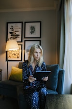 A person wearing dark blue pajamas with white star patterns is sitting on a comfortable sofa, holding a tablet. The room is softly lit by a nearby floor lamp, with framed artwork adorning the wall behind them. A green cushion adds a touch of color to the sofa.