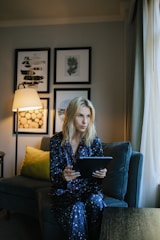 A person wearing dark blue pajamas with white star patterns is sitting on a comfortable sofa, holding a tablet. The room is softly lit by a nearby floor lamp, with framed artwork adorning the wall behind them. A green cushion adds a touch of color to the sofa.