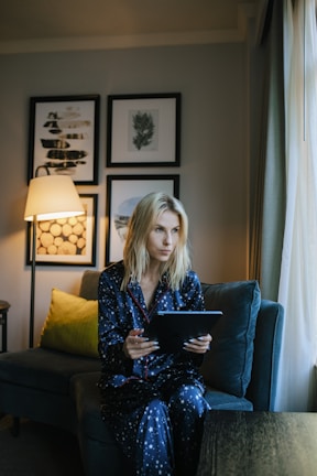 A person wearing dark blue pajamas with white star patterns is sitting on a comfortable sofa, holding a tablet. The room is softly lit by a nearby floor lamp, with framed artwork adorning the wall behind them. A green cushion adds a touch of color to the sofa.