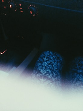 An interior view of a vehicle, featuring a patterned textile, possibly clothing, in the foreground. The dashboard is dimly lit with red indicators and dials, creating a low-light environment.