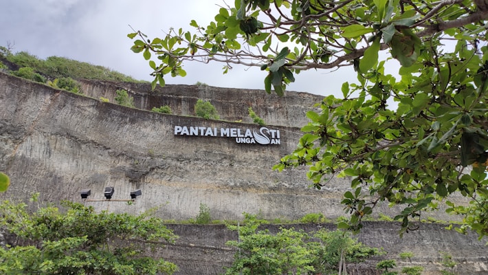 A rocky cliff with signage reading 'Pantai Melasti Ungasan' is partially obscured by lush green foliage. The cliff features layered stone formations, and there's a cloudy sky overhead.