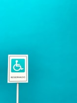 A vertical sign with a white background and a blue symbol of a person in a wheelchair, indicating accessibility, with the word 'Reservado' written below. The sign is set against a solid turquoise blue background.
