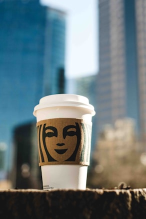 A takeaway coffee cup with a white lid is adorned with a cardboard sleeve featuring a stylized face design. The cup is placed outdoors with a blurred backdrop of modern high-rise buildings, indicating an urban setting.