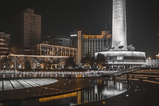 A night scene of a modern urban area featuring a tall, illuminated tower with large satellite dishes on its lower level. Surrounding the tower are multi-story buildings with a mix of traditional and modern architectural styles. The area includes landscaped grounds with water features, such as pools and reflective surfaces, lined with lights that add a warm glow to the surroundings.