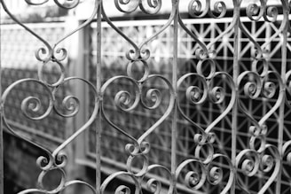 An intricately designed wrought iron gate featuring numerous elegant heart-shaped patterns and scrollwork. The gate appears weathered, revealing a sense of age and artistry. In the background, there is a blurred fence that complements the ornate design of the gate.