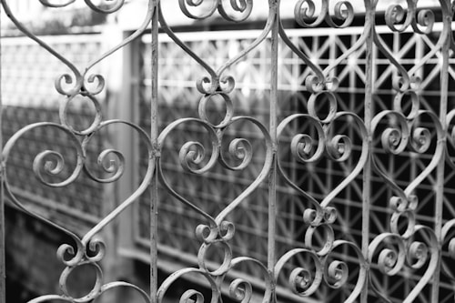 An intricately designed wrought iron gate featuring numerous elegant heart-shaped patterns and scrollwork. The gate appears weathered, revealing a sense of age and artistry. In the background, there is a blurred fence that complements the ornate design of the gate.